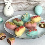 Bunte Dattel-Ostereier auf einem Teller – gefüllt mit Nussmus und umhüllt von farbiger veganer Schokolade, dekoriert mit Nüssen und gefriergetrockneten Beeren.