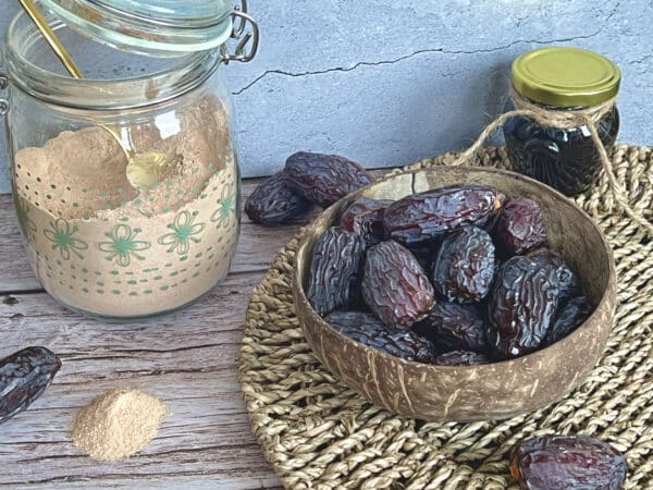 Dieses Bild zeigt hochwertige Medjool-Datteln in einer Schale, daneben ein Glas mit Dattelzucker und ein kleines Gefäß mit Dattelsirup. Die Aufnahme wurde auf einem rustikalen Holztisch mit natürlichem Licht aufgenommen und symbolisiert die Vielfalt natürlicher Süßungsmittel als gesunde Alternative zu raffiniertem Zucker.